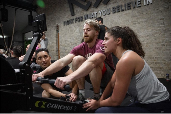 Personal training session with man leaning forward on rowing machine with another man and personal trainer holding his feet